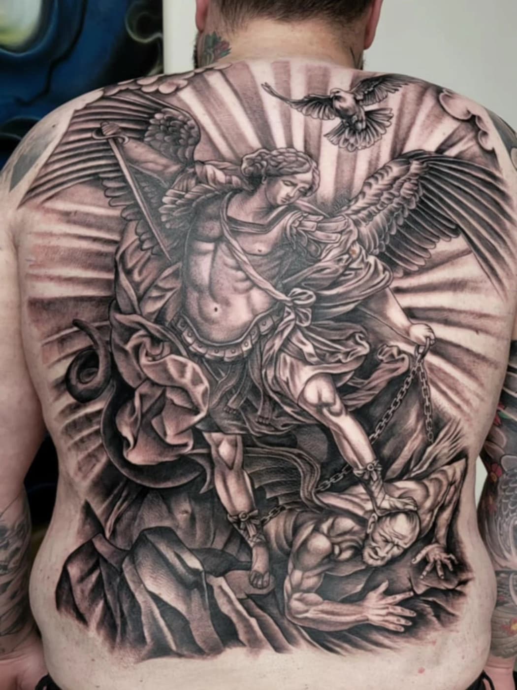 Full back piece: a winged archangel with a sword standing over a chained figure, rays and a dove above