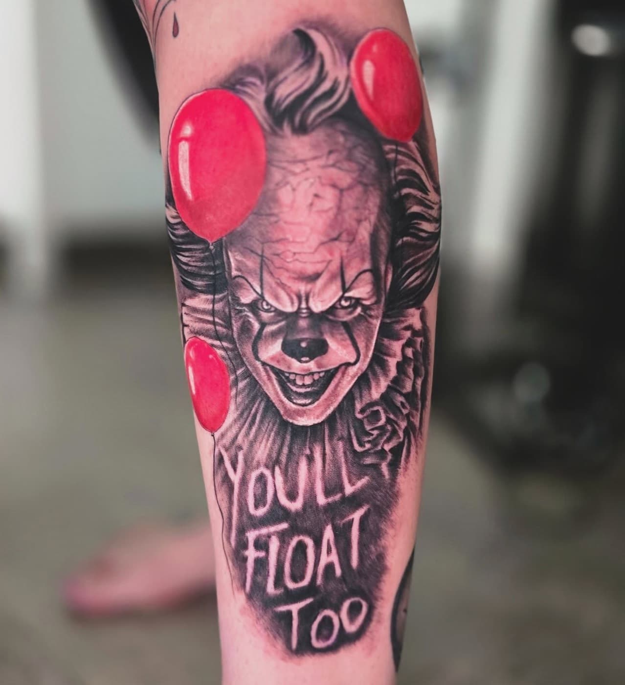 Horror-film clown with red balloons and lettering reading “you’ll float too”, on a lower leg