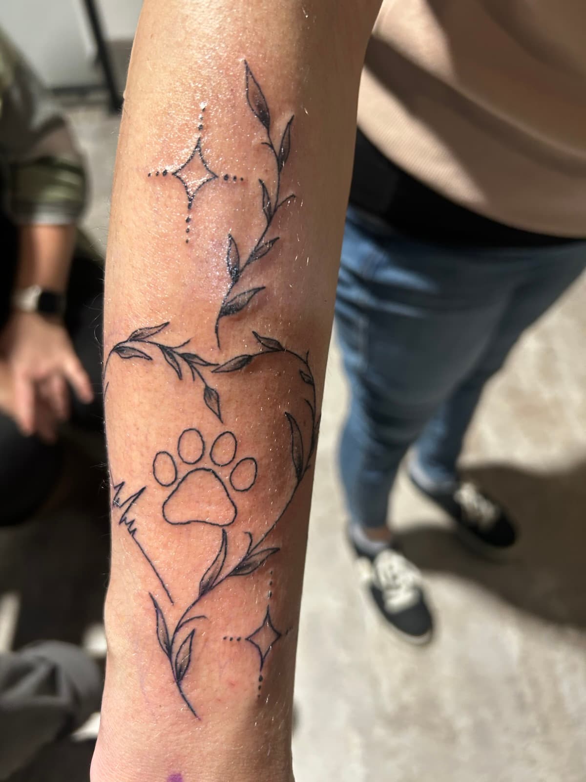 Paw print inside a heart of leafy vines with a heartbeat line, fine black line on the forearm
