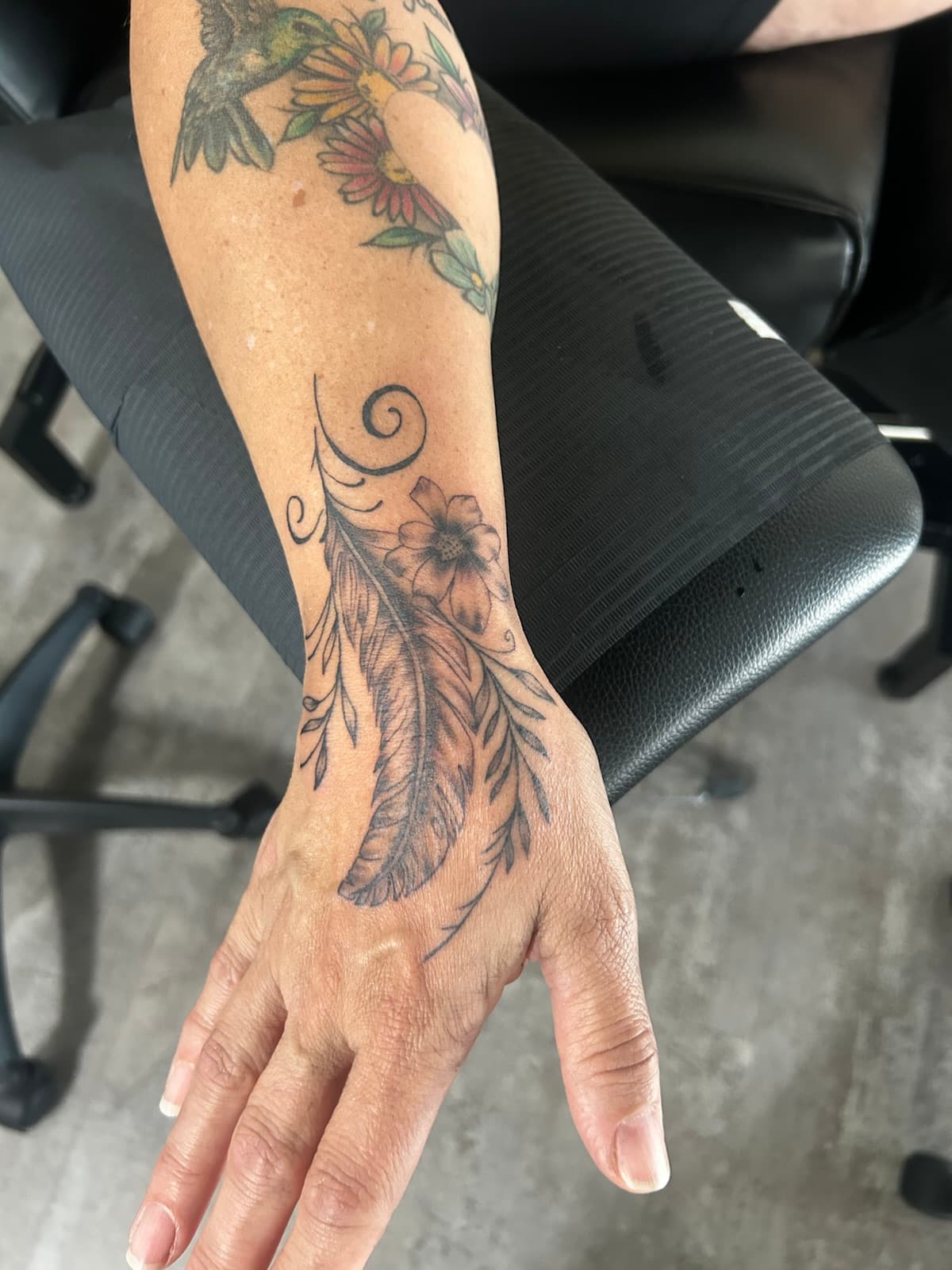 Feather with a flower and swirling vines running onto the hand, black and grey on the wrist