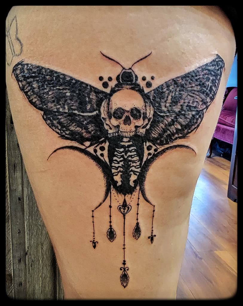 Death’s head moth with a skull on its back and hanging jewelled chains, black and grey on the thigh