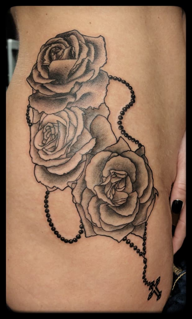 Three roses wrapped in rosary beads ending in a cross, black and grey on the side of the torso