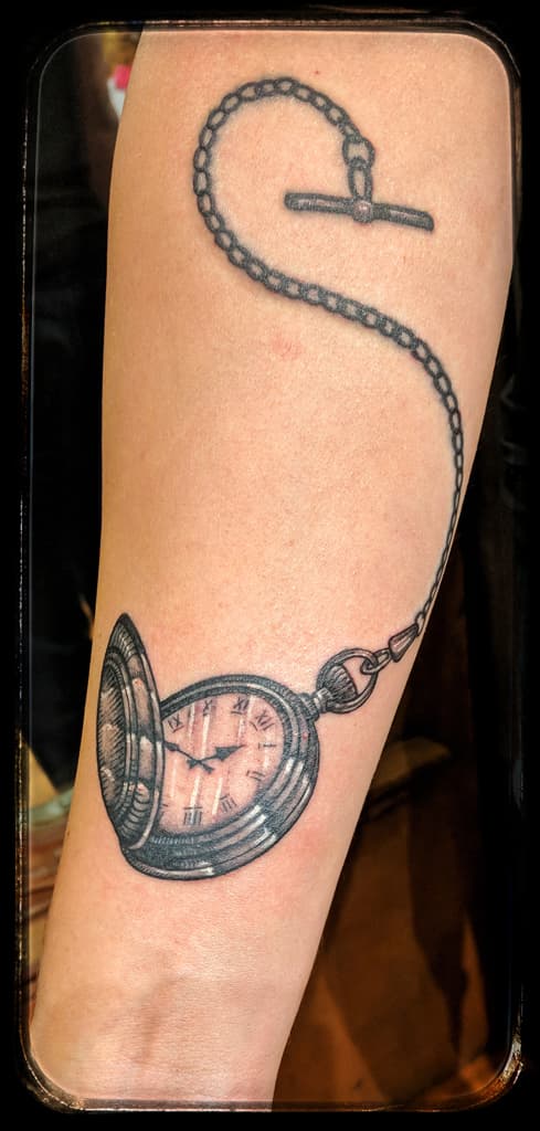 Open pocket watch with Roman numerals, its chain looping up to a T-bar, black and grey on the forearm