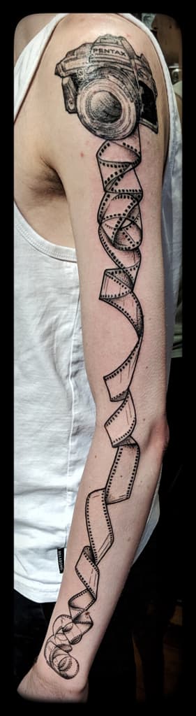 Pentax film camera on the shoulder with a film strip spiralling down to the wrist, black and grey full arm