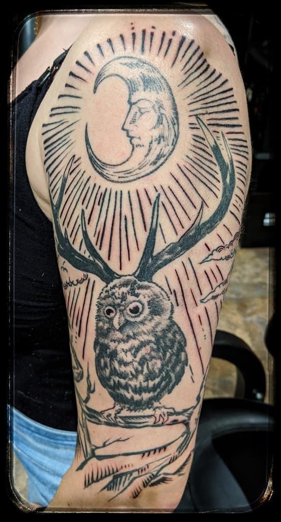 Antlered owl perched on a branch under a crescent moon with a face and radiating lines, blackwork on the upper arm