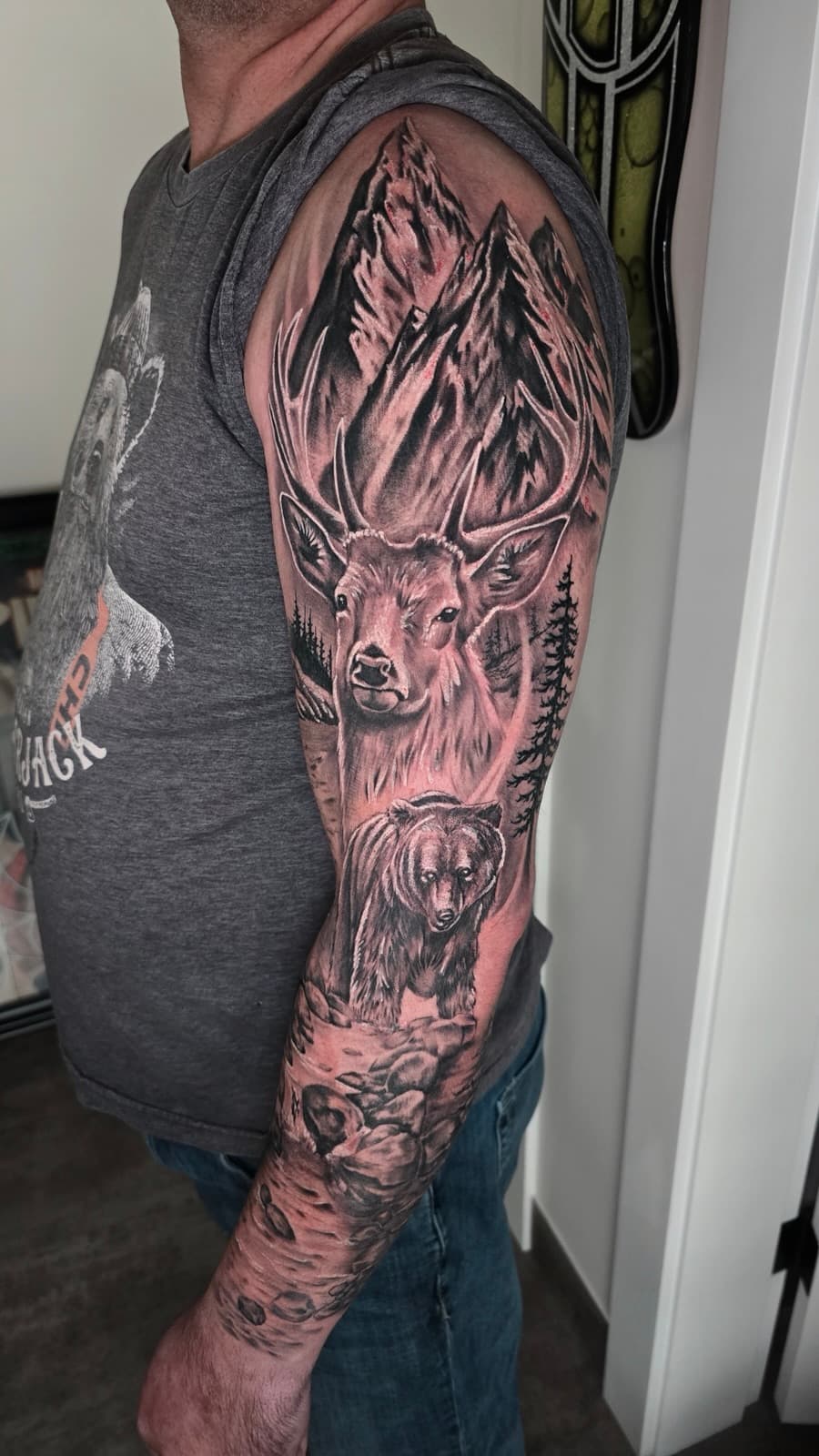 Stag in a pine forest under mountain peaks with a bear below, black and grey sleeve from shoulder to wrist