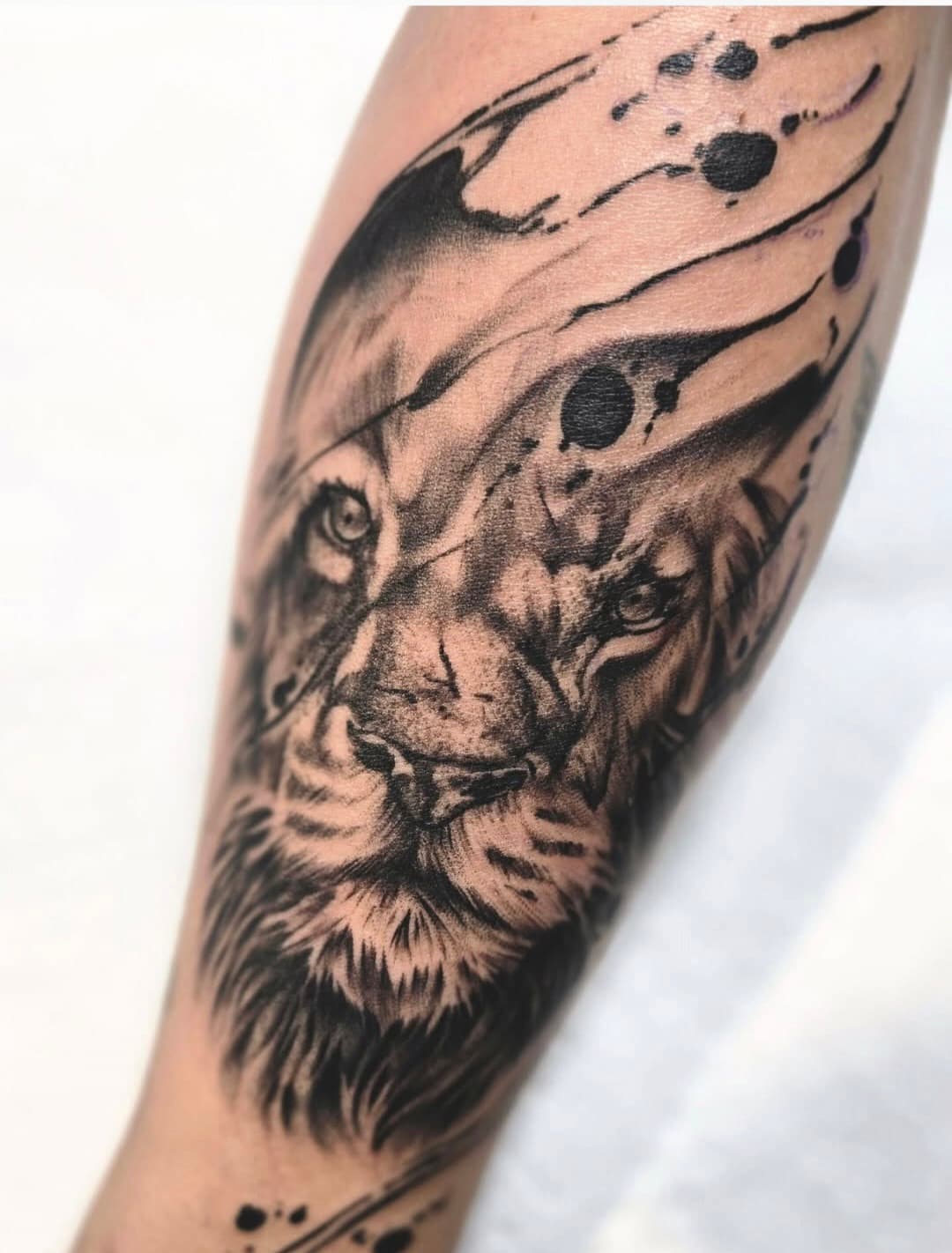 Lion’s face emerging from black ink splatter, black and grey on the forearm